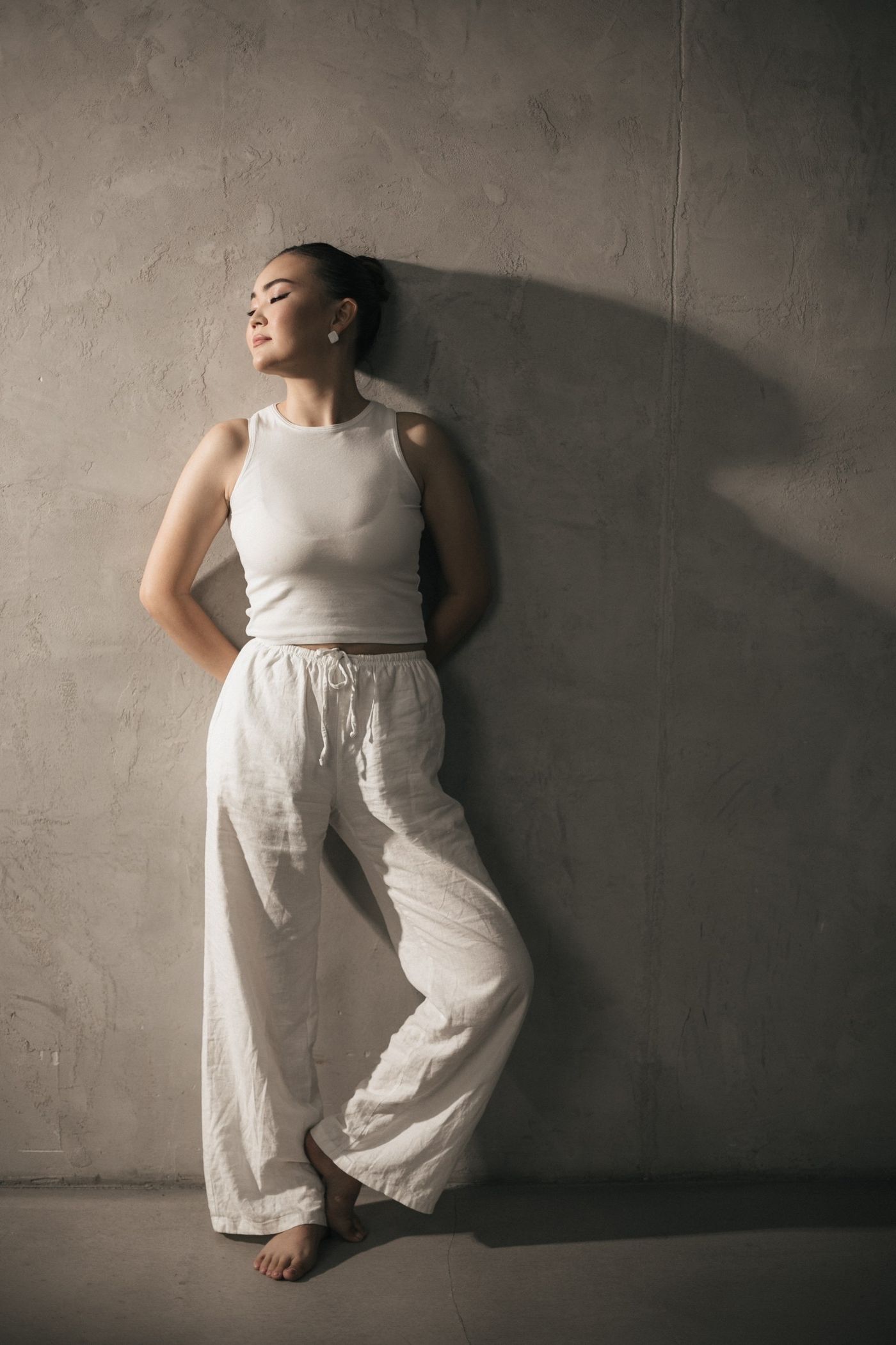 Woman in pale linen trousers and a sleeveless white top against a textured wall.