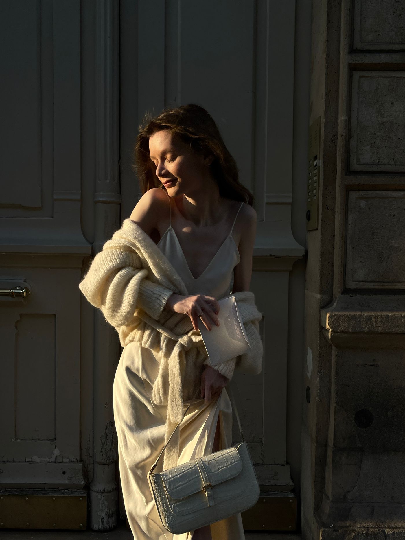 Woman in a cream outfit with a cardigan and small shoulder bag at dusk.