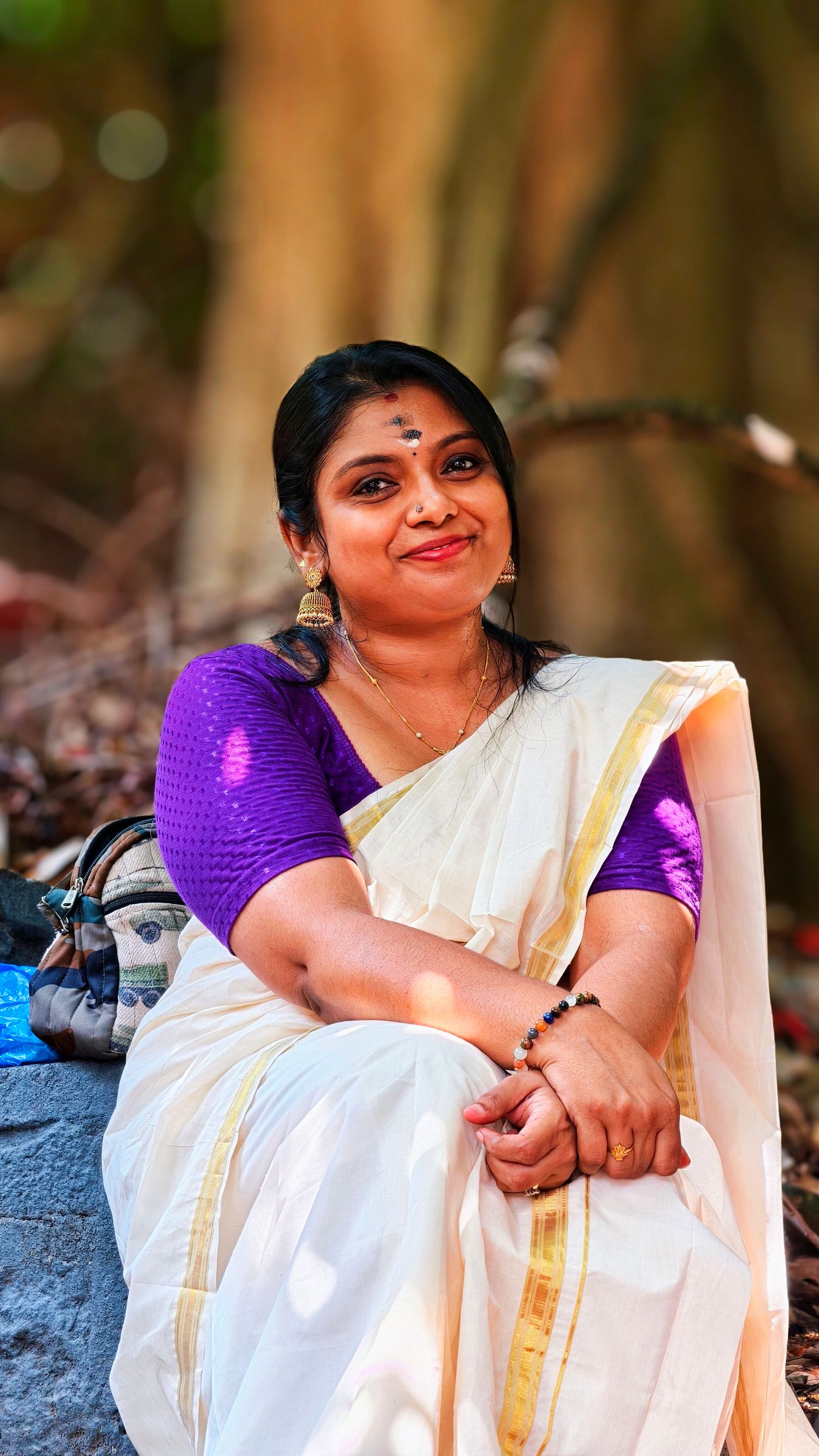 Seated woman wearing a white and gold Kerala saree with a purple blouse.