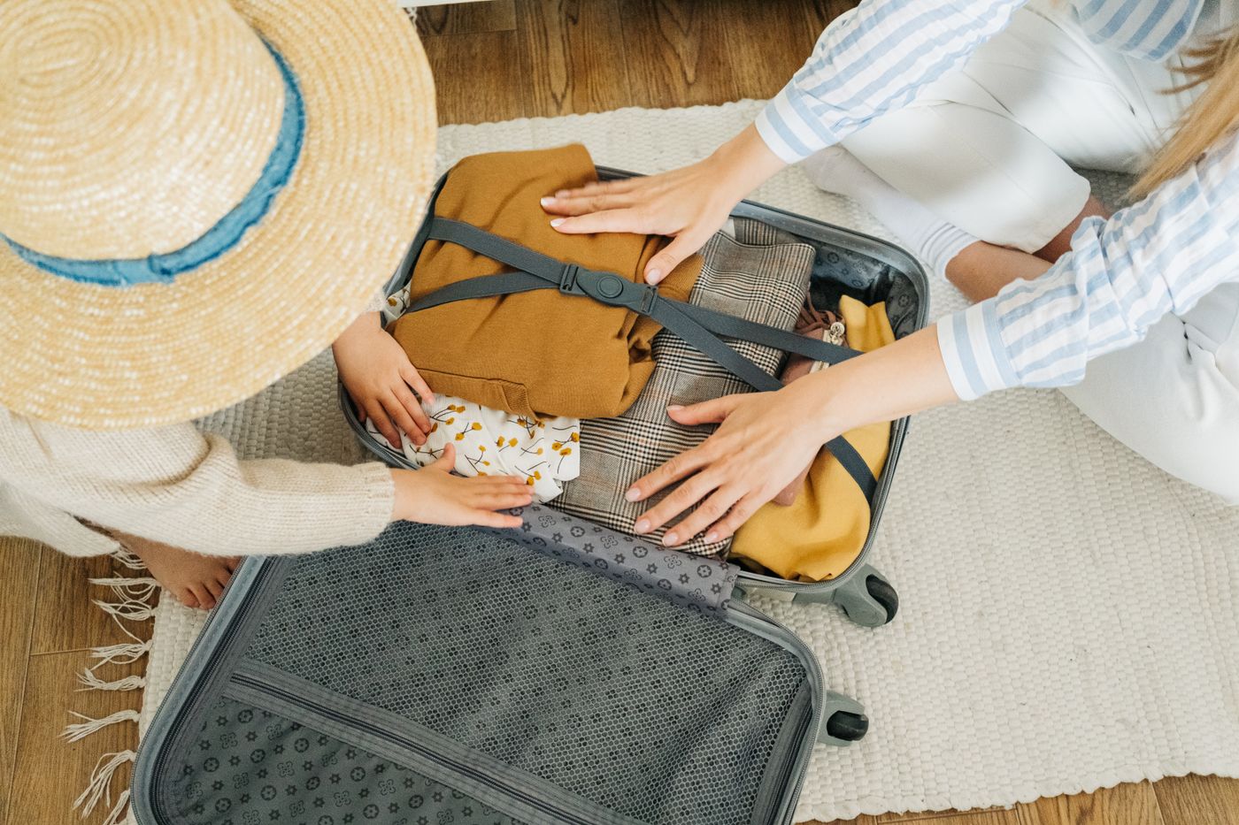 Hands arranging light-coloured holiday clothing in a suitcase beside a straw hat.
