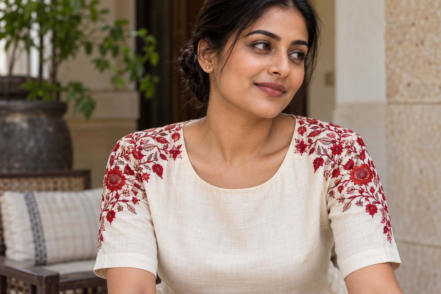 Woman wearing tiny stud earrings with a cream blouse embroidered in red across both shoulders.