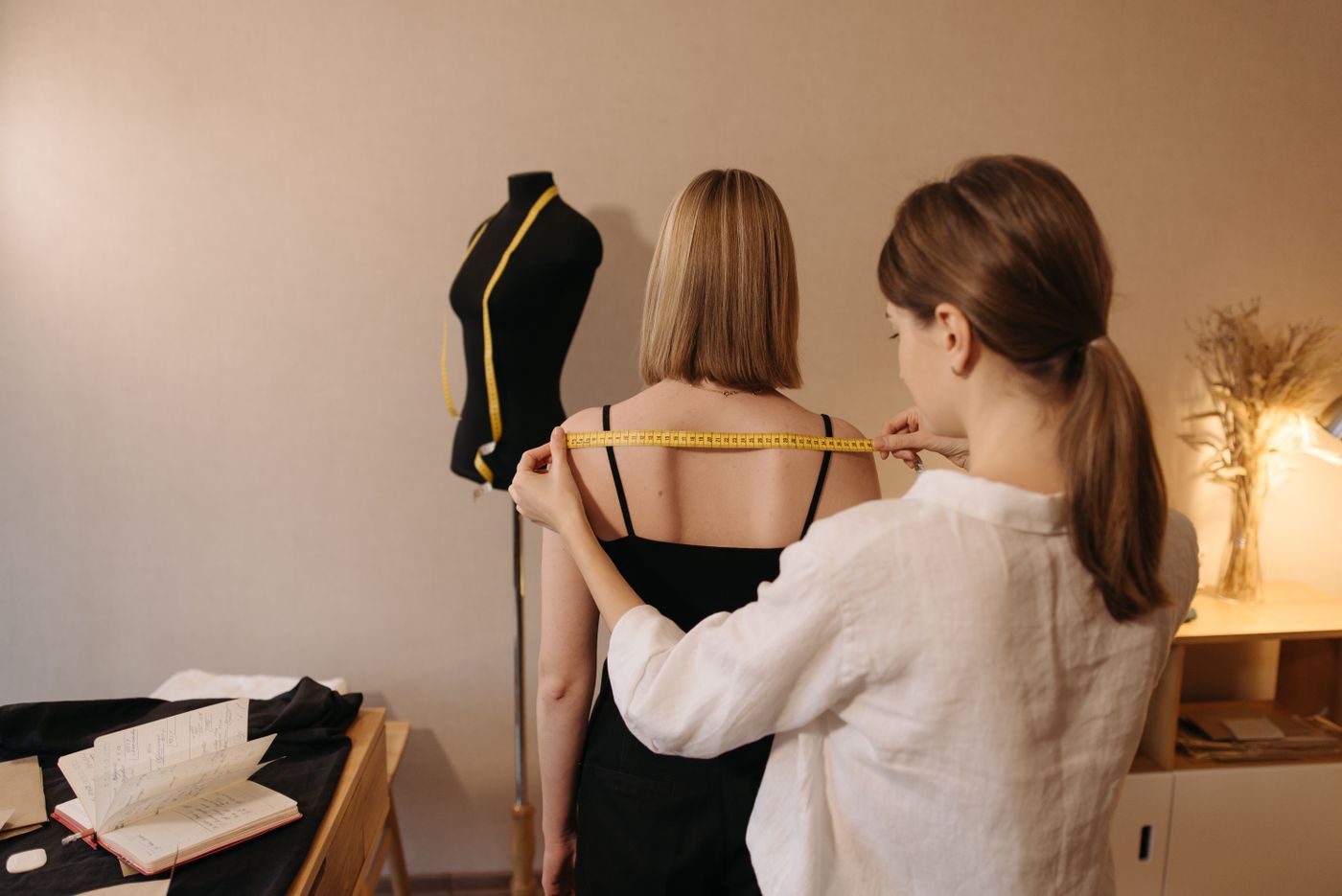 Tailor measuring the shoulder width of a woman in a dark top.